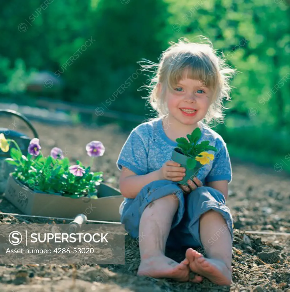 Happy little blonde girl sits beside a box of purple pansies in a garden and holds a small pot containing a yellow pansy.