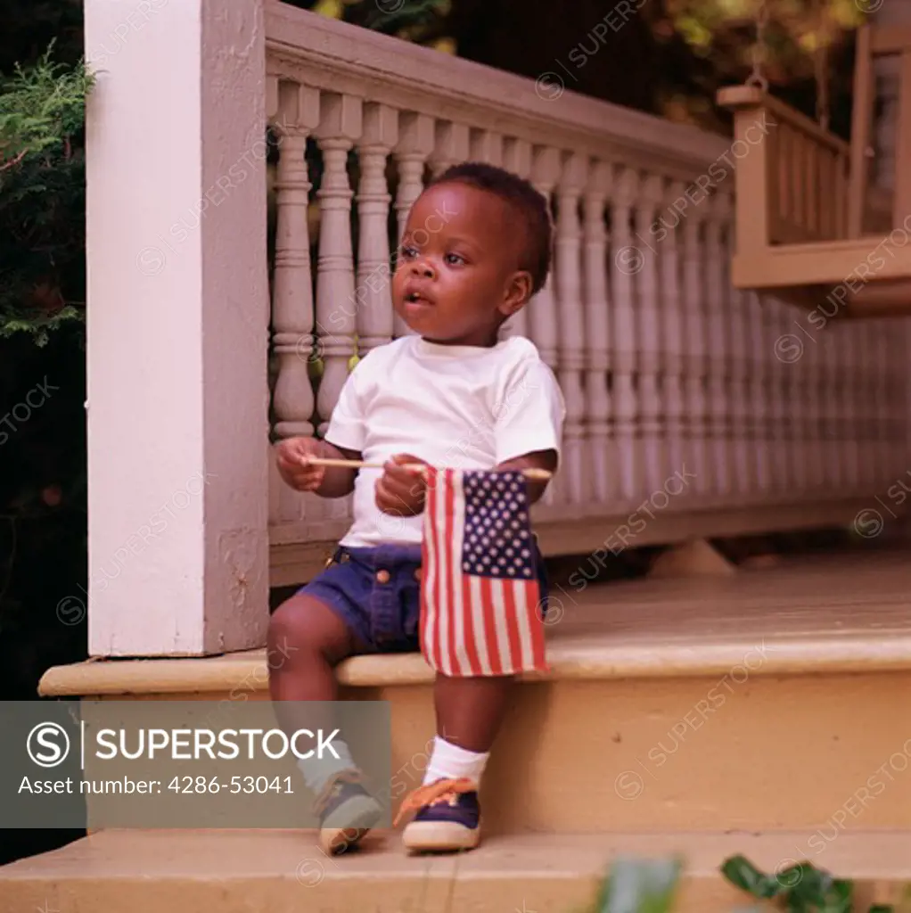 An African-American toddler sitting on the steps of the front porch holding a small American flag. 