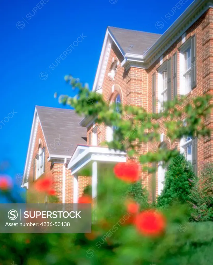 Exterior angled view of front of two story red brick house partially obscured by red flowers in foreground. Property released.