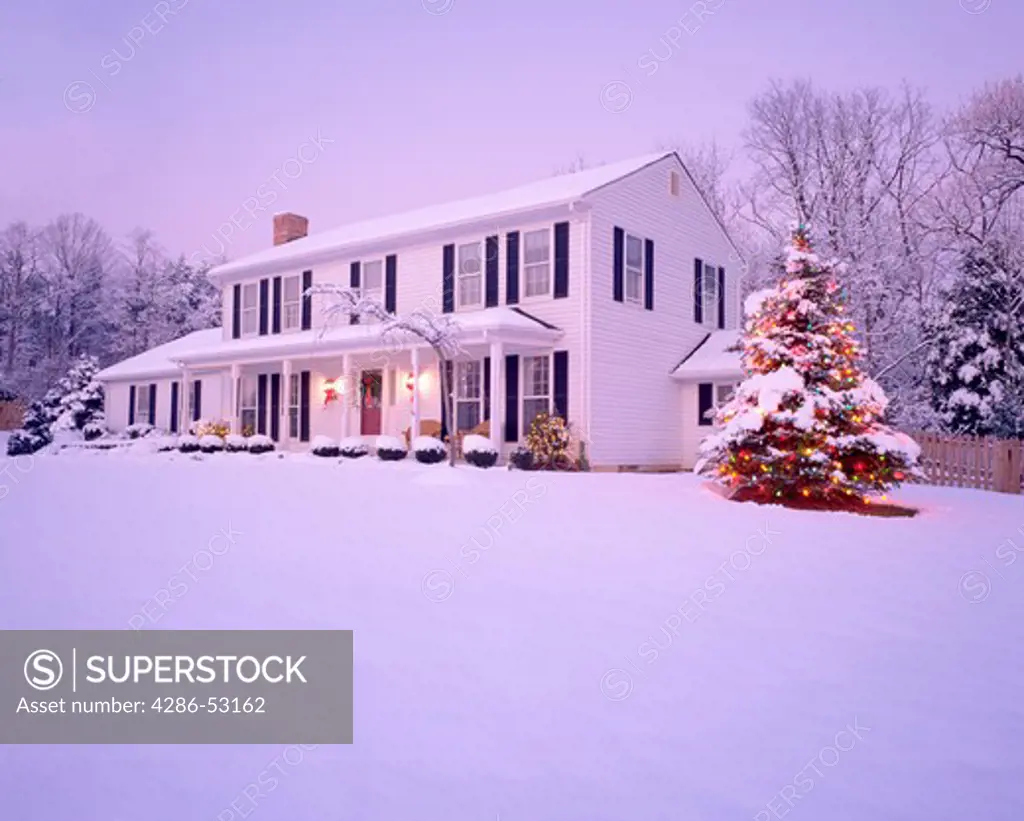 Snowy winter view of exterior of two story white frame house with front porch and tree lit with Christmas lights.  Property released.