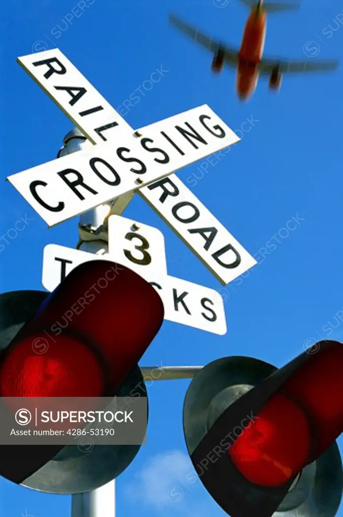 Close-up of a railroad crossing sign and lights while a jetliner flies low above.