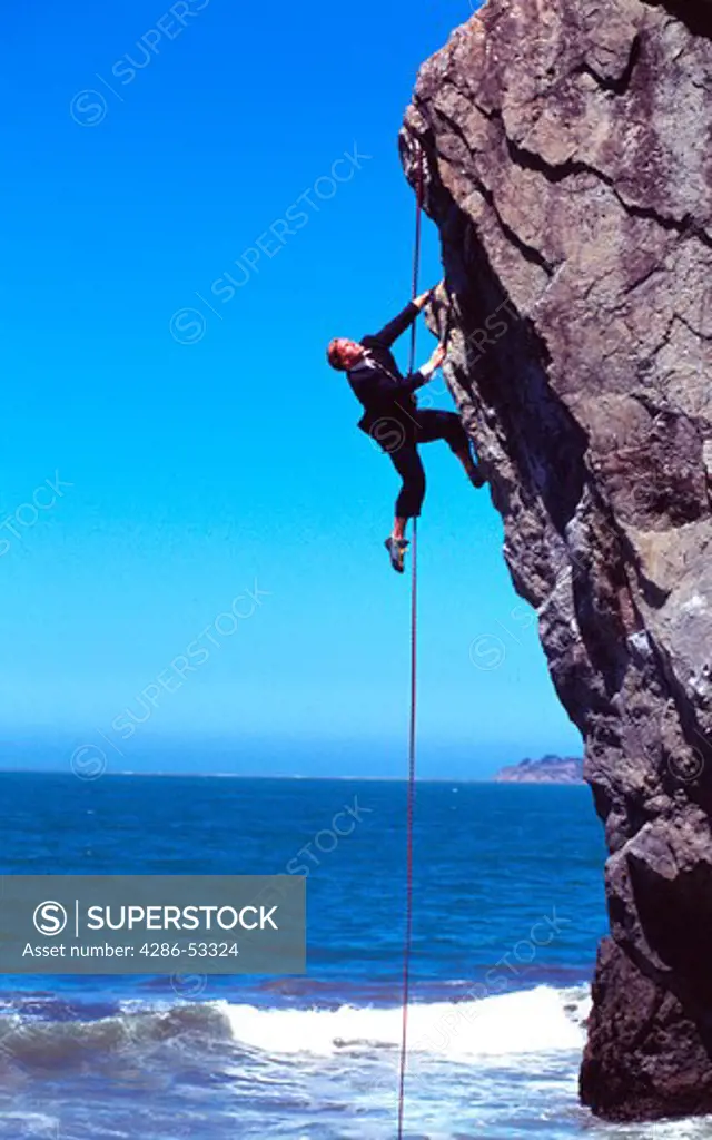 Businessman climbing up rock face over ocean.