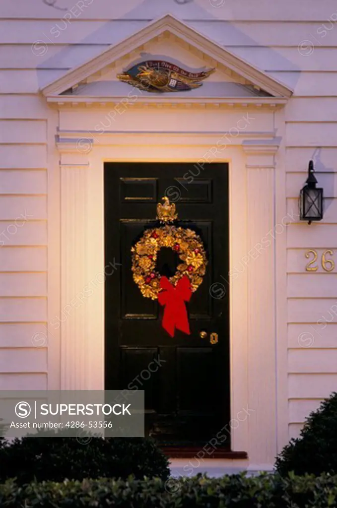 Spot lit gilded pine cone wreath with a red bow hanging on a black front door to a white frame house.