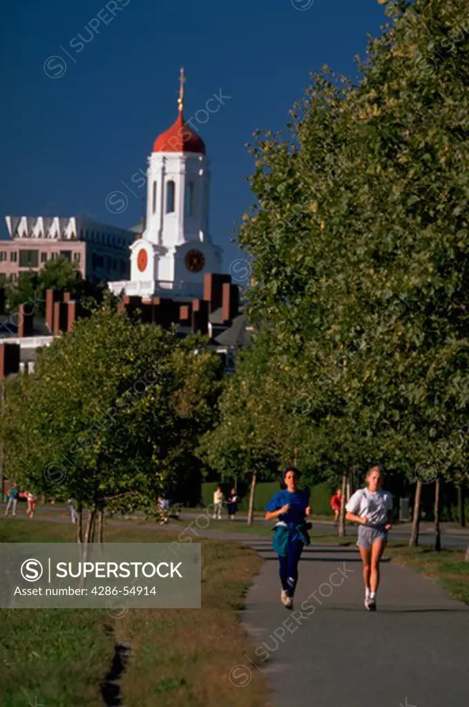 Two women jog along path on banks of Charles River with Harvard University's Dunster House at rear, Cambridge, Massachusetts.