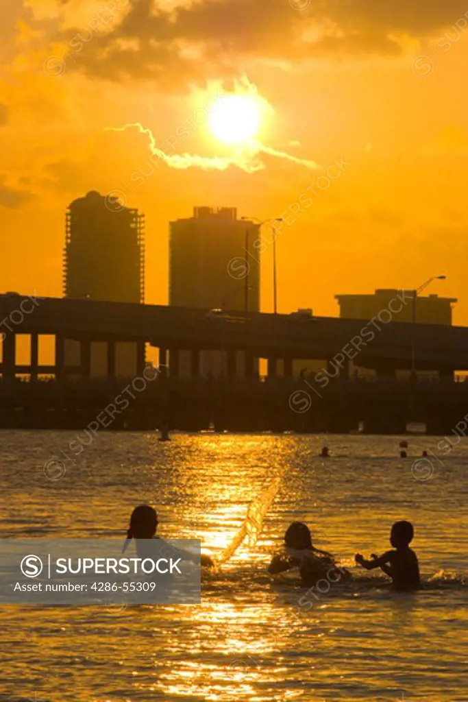 MIAMI, FLORIDA, USA - Swimmers play in shallow water, as sun sets over Biscayne Bay, with buildings and causeway at rear.