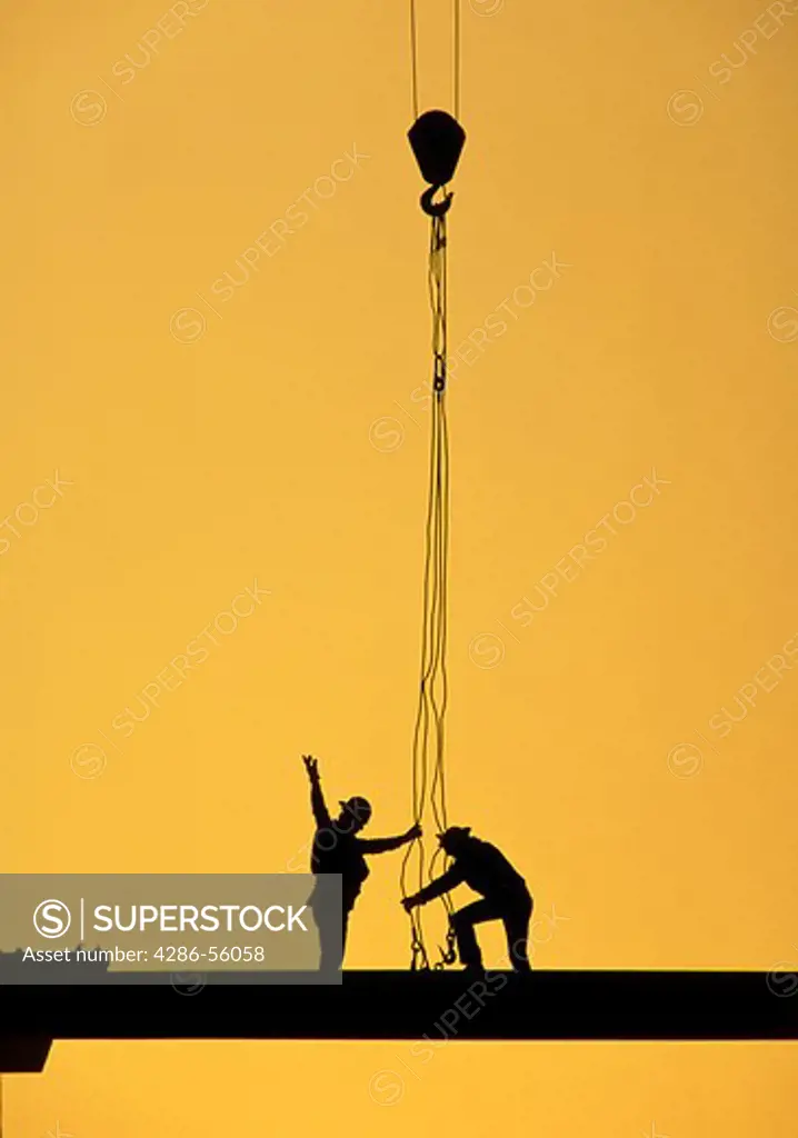 Silhouette of two construction workers standing on a beam as a crane lowers grappling hooks.