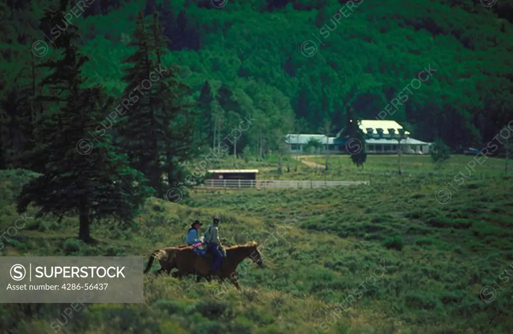 Two men riding horse back with a ranch house in the background.