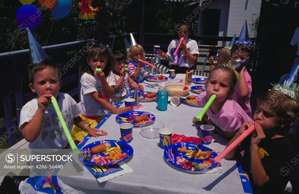 Nine children with noise makers and party hats sitting around a table outside at a birthday party.