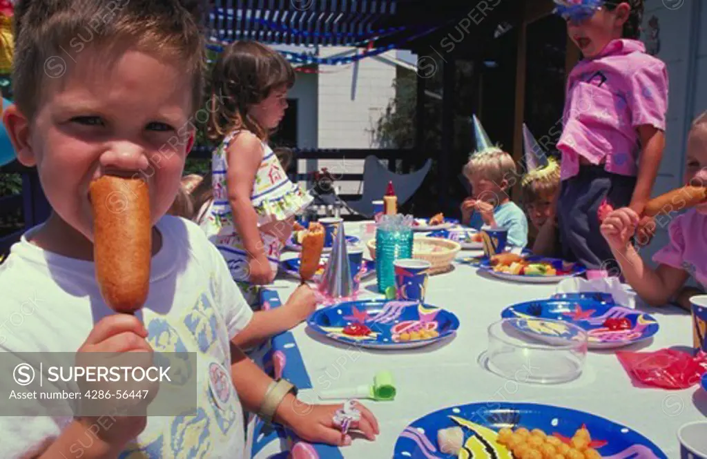 Portrait of a young boy eating a corn dog while other children play at a birthday party in the background.