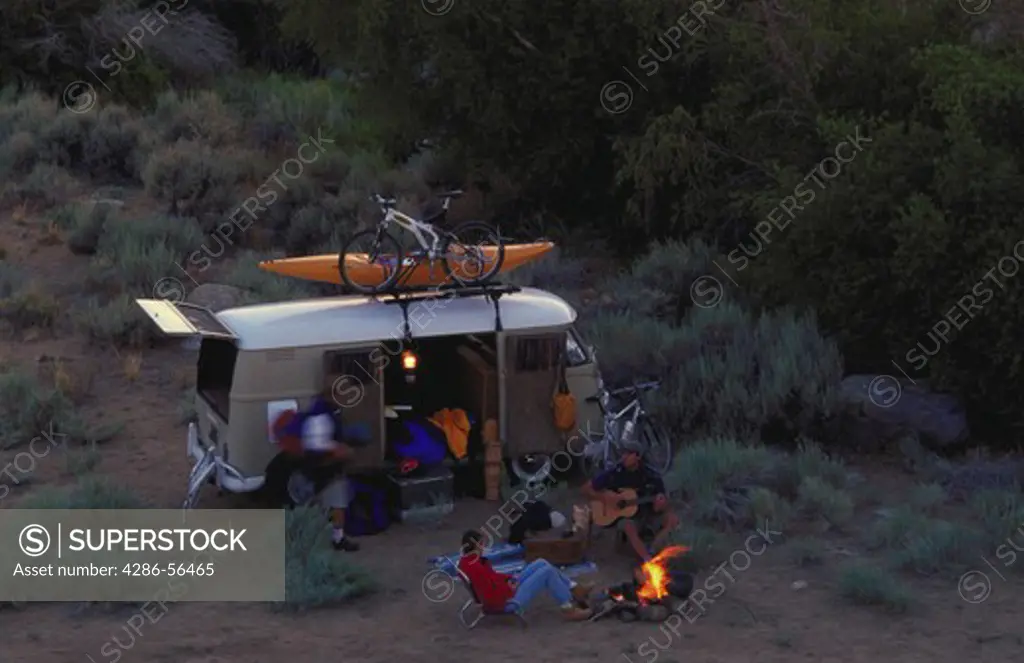 Aerial view of two men and a woman around a campfire next to their van. The have mountain bikes and a kayak with them. There are mountains in the background.