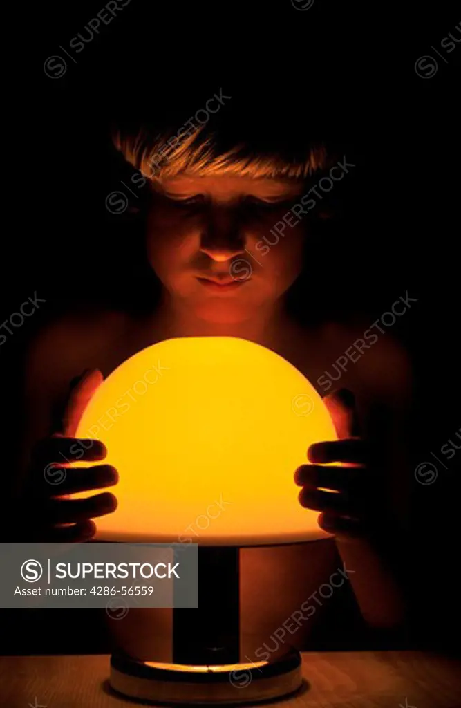 Young boy in shadows looking at electric light while holding lightshade.
