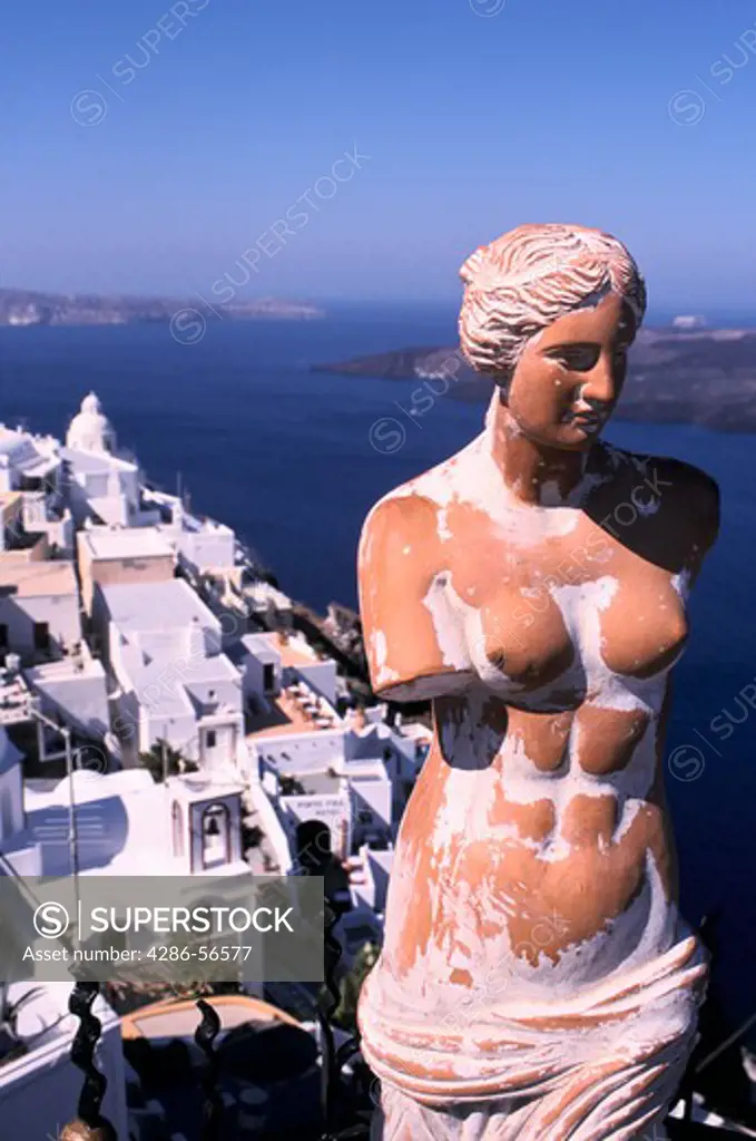 Terra cotta statue of Aphrodite with faded whitewash stands before white buildings of Santorini Greece and deep blue Agean Sea.