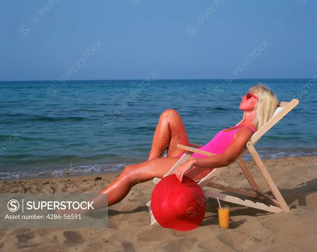 Blonde woman in pink bathing suit sitting on a deckchair on the beach and holding a large red sun hat.