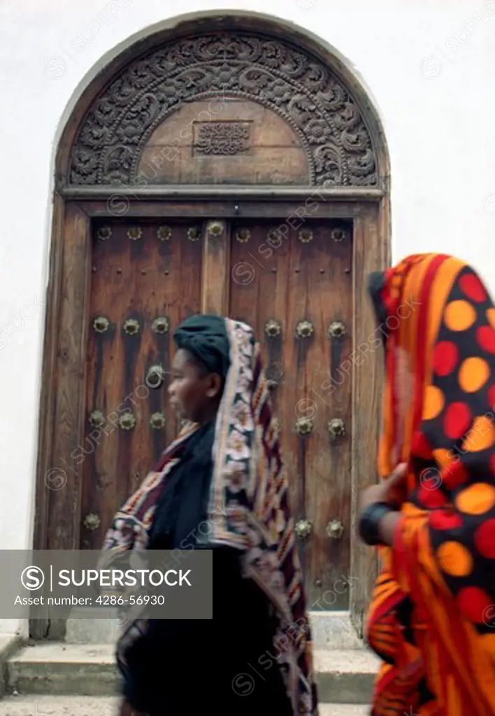 Two Zanzibari women in traditional khanga robes, walking past a magnificent carved wooden door. Stone Town. Zanzibar. Tanzania. East Africa.