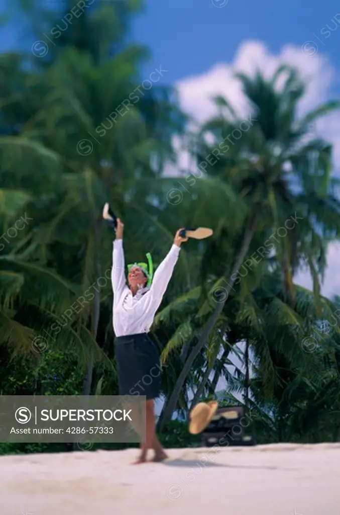 Businesswoman in skirt, white blouse and green snorkel tosses her shoes overhead on a sunny, sandy beach of the Maldives Islands.