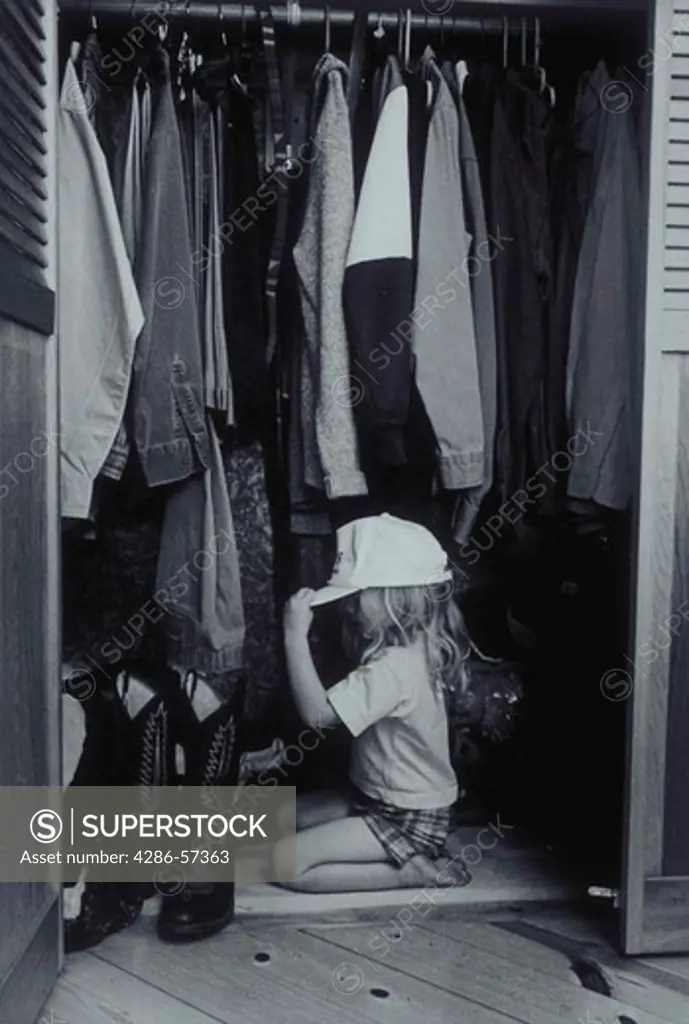 Black and white image of a young girl kneeling on her fathers closet floor trying on her dads hat.