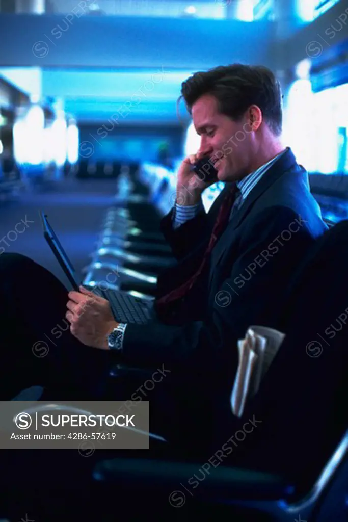 A businessman sitting in an airport terminal talking on a cell phone with his laptop computer open on his lap.