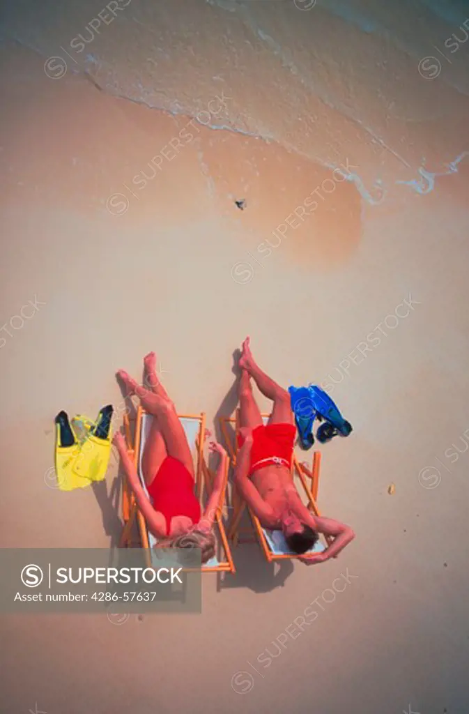 Aerial view of a couple wearing bathing suits sitting in chairs on the beach at the edge of the water with their snorkel masks and fins along side them.