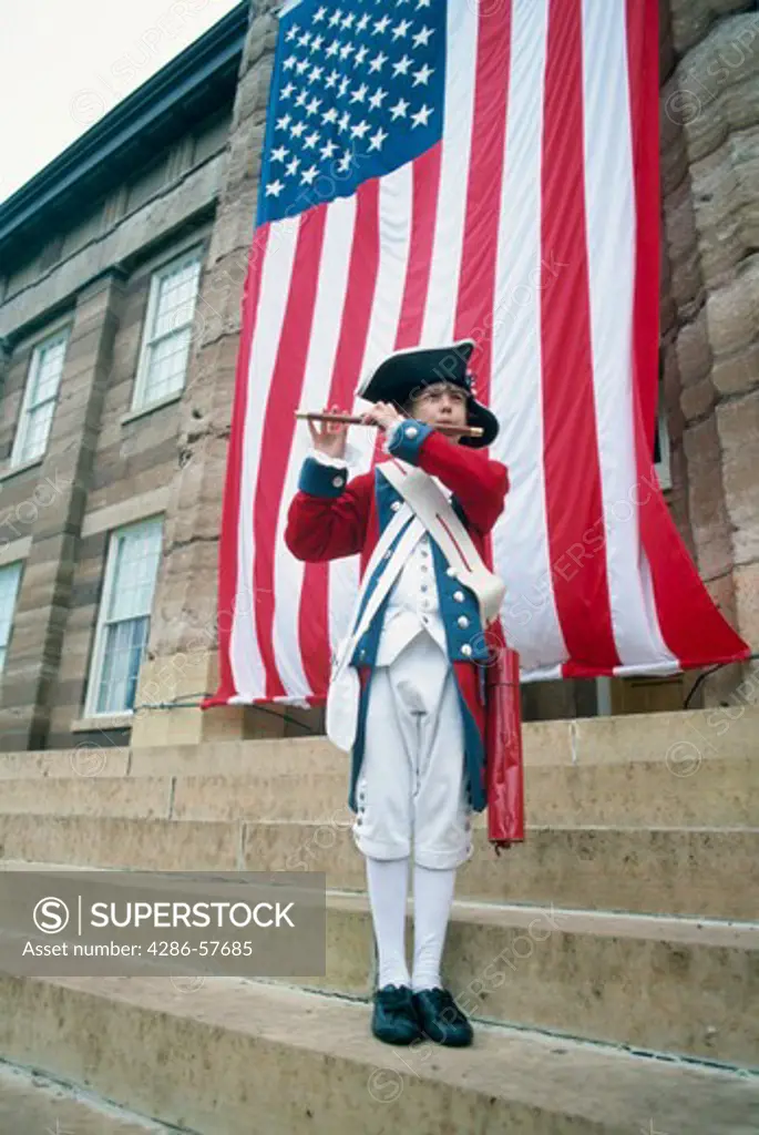 Man dressed in coloniel garments in front of a large United States flag playing a fife on the 4th of July.