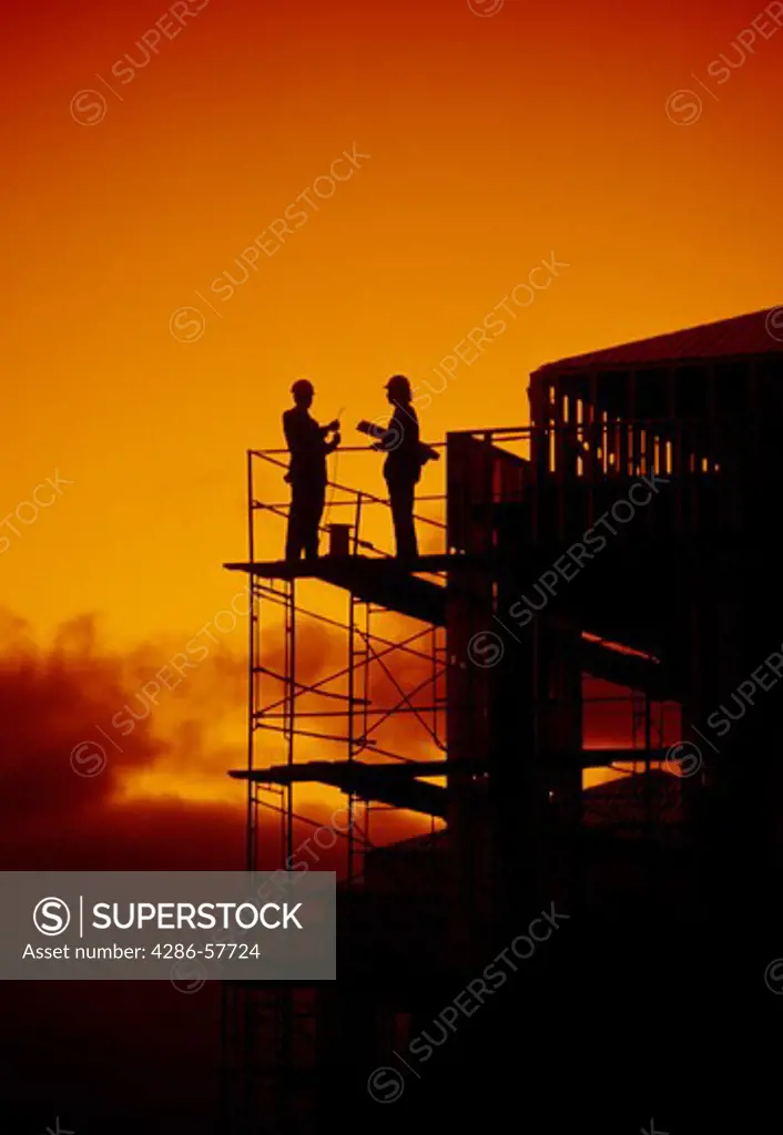 Two men silhouetted against an orange sky stand high above the ground on a platform discussing plans for a building under construction.