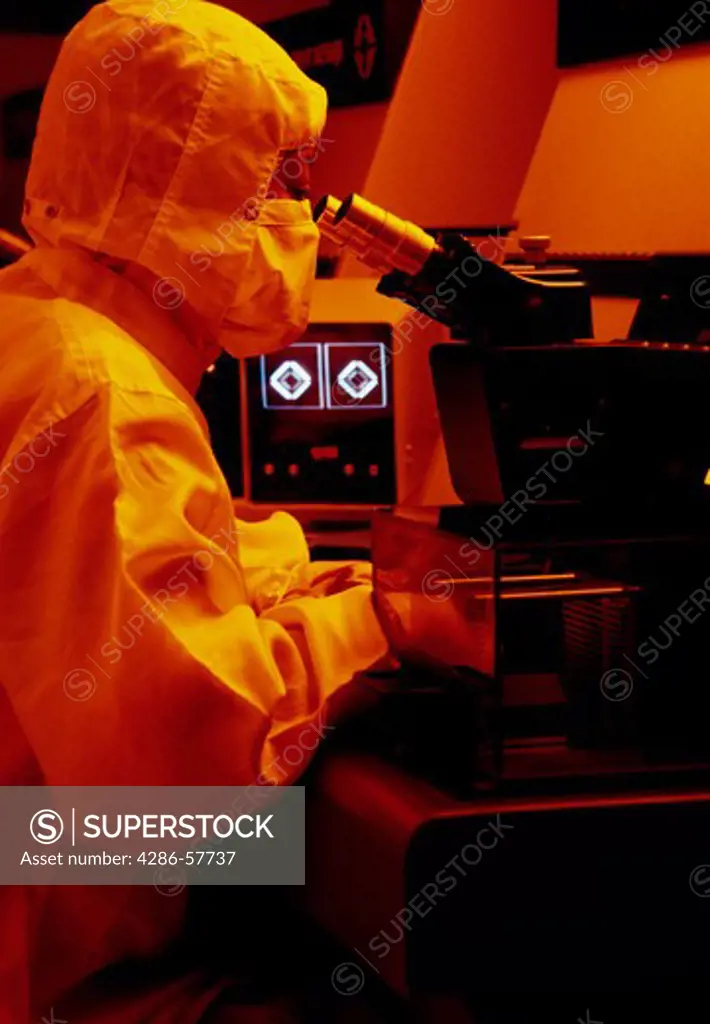 A lab technician wearing protective gear examining an item under a microscope in the lab. 