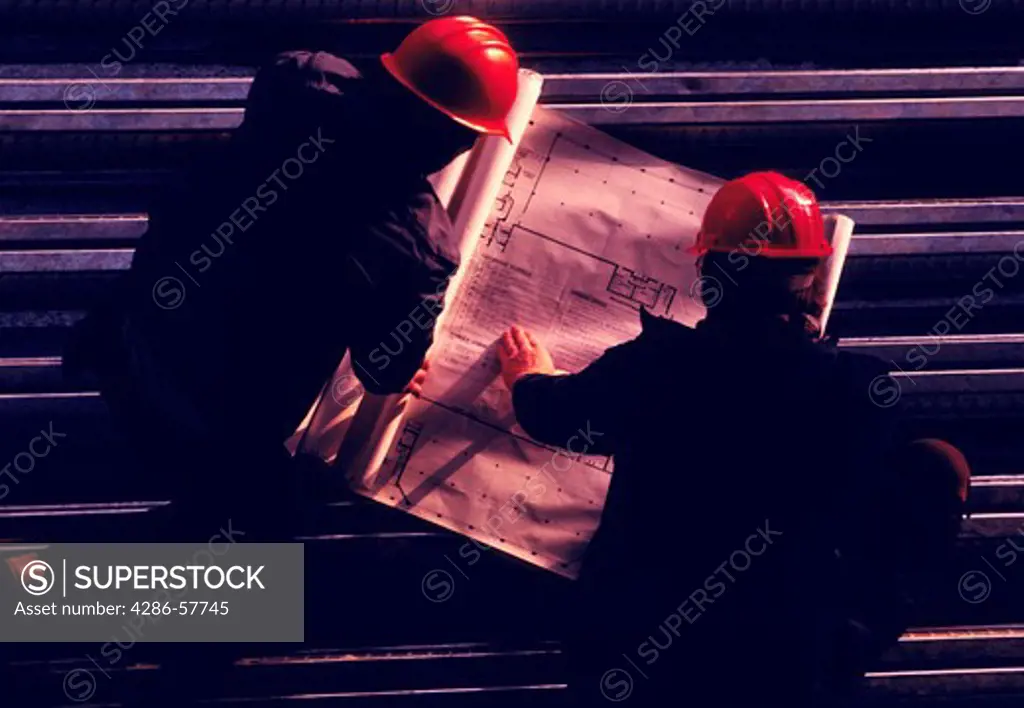 Birds eye view of two construction workers wearing hard hats looking over blue prints.
