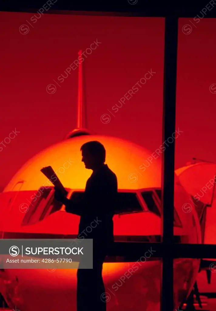 A business man reading a newspaper standing in an airport terminal with the commercial airliner insight through the windows.
