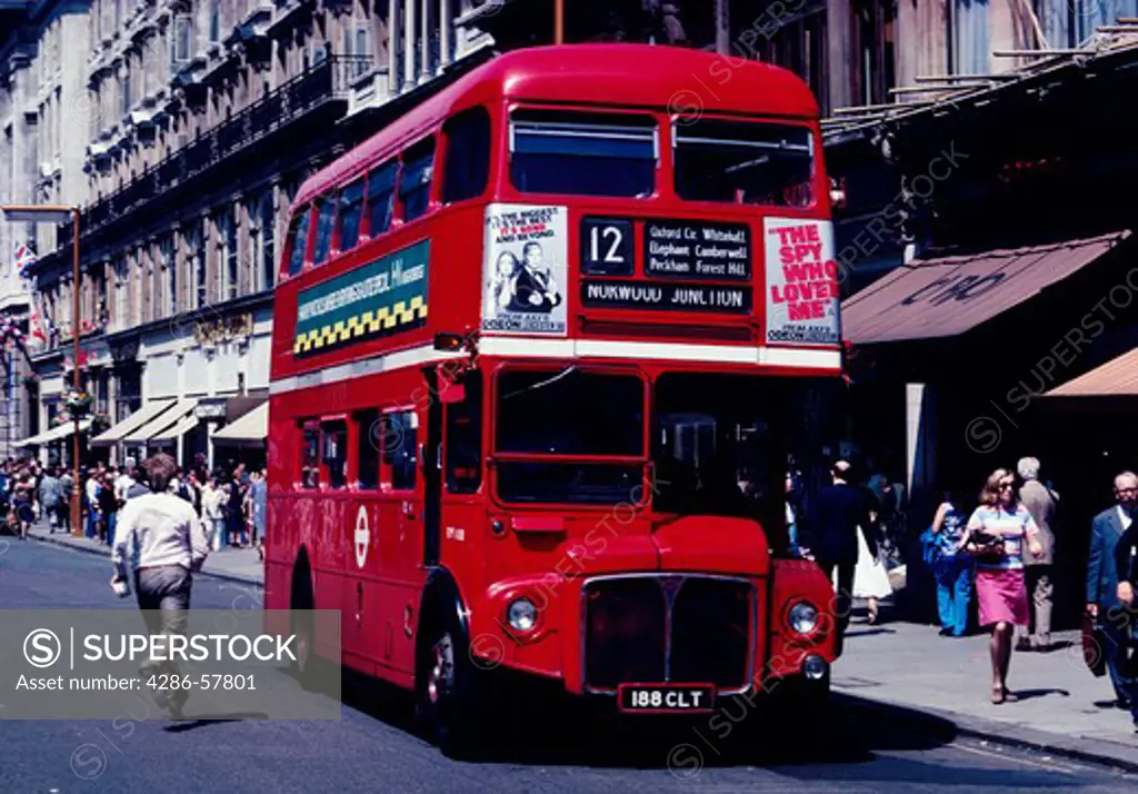 A large, red double decker bus in the city streets of London, England with several pedestrians near the bus.