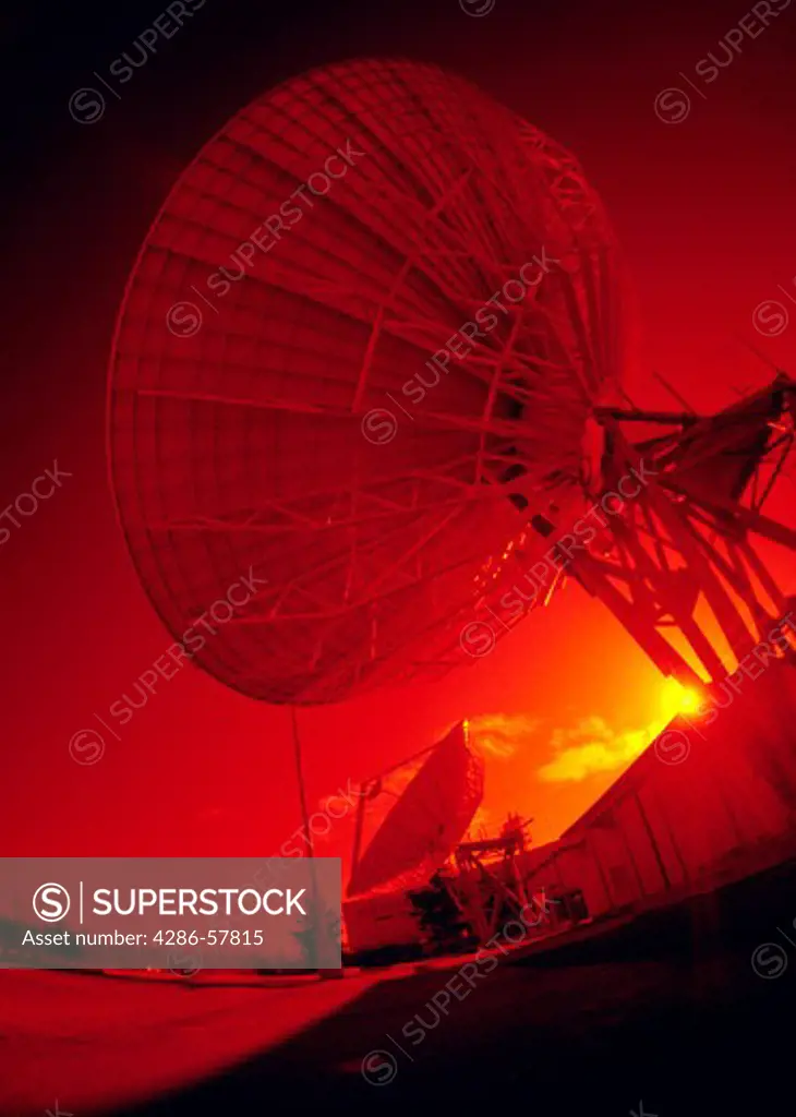 Side-view of a large, communication microwave dish with the sun setting behind it.