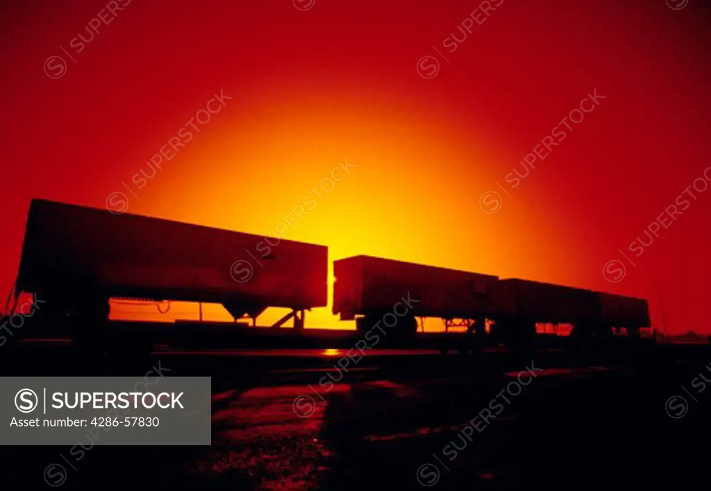 Side view of piggy back trailers on rail cars with the sun setting in the background.