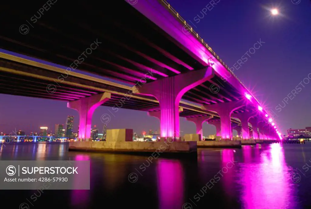A moon shines down at night, just before sunrise, on Rickenbaker Causeway bridge over Biscayne Bay in Miami, Florida as magenta lights illuminate the bridge as part of a beautification project.