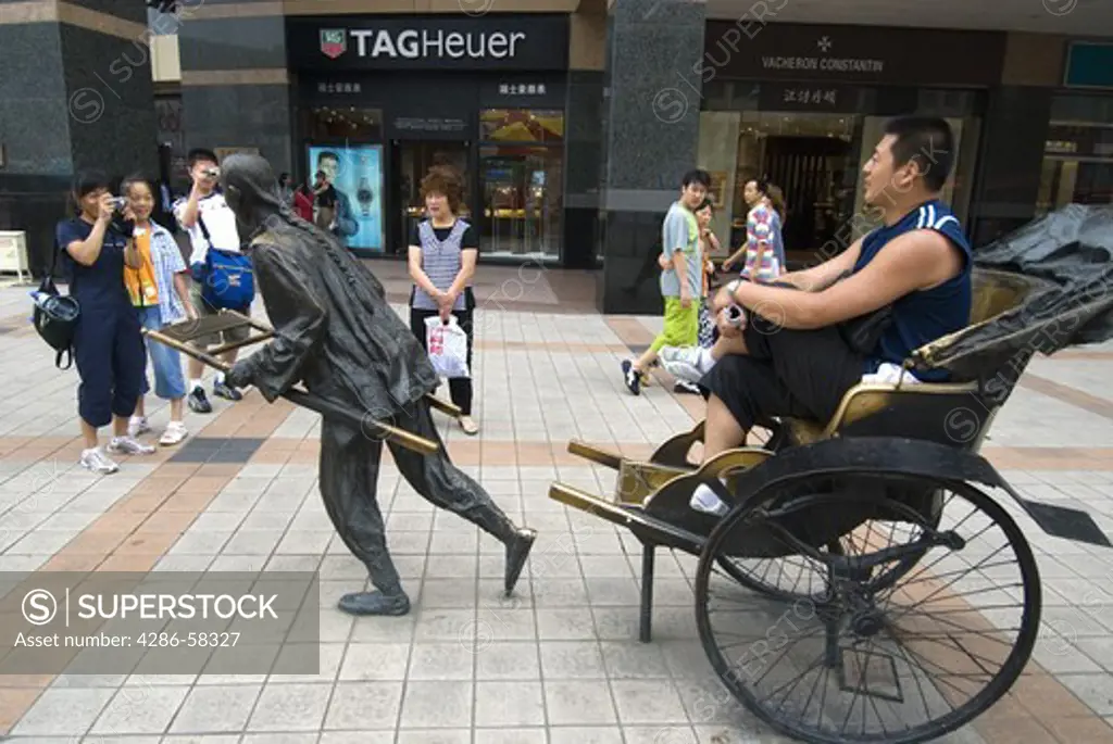 Teenagers photograph adult posing with sculpture of rickshaw on Wang Fujing Street, a high end shopping district,  Beijing, China.