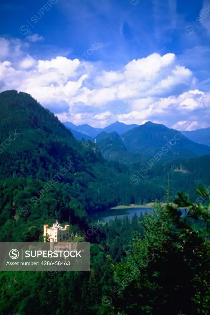 View of King Ludwigs Homenschwangau Castle, Bavaria, Germany  with lush green mountains, countryside, and blue sky with clouds in the background.