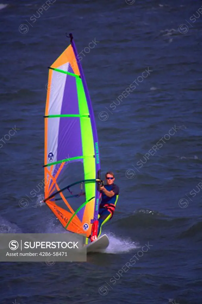 A man in a wet suit riding on a wind surfer in the ocean. The wind surfer uses wind and a sail for power.