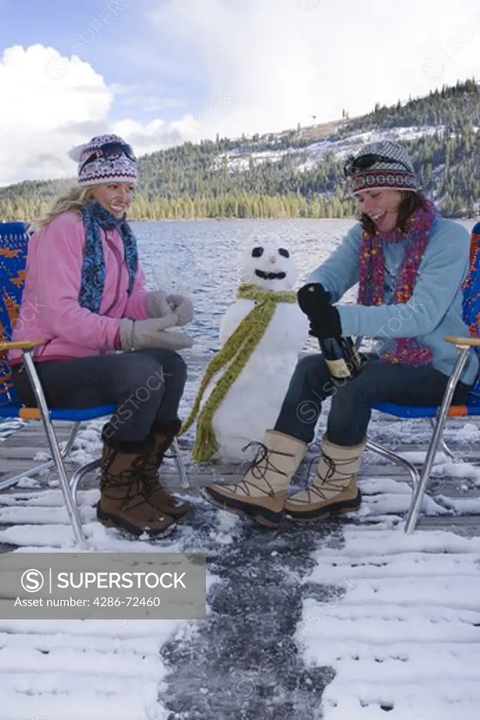 Two women friends drinking champagne on a dock on a lake with a snowman on a cold winters day in Truckee, California