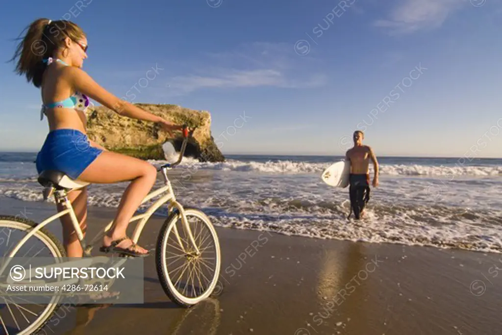 A woman on a cruiser bike and a man with a surfboard on the beach at Natural Bridges State Park in Santa Cruz in California