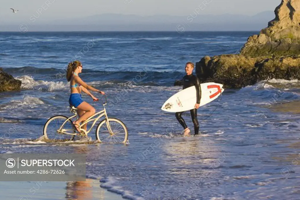 A woman on a cruiser bike and a man with a surfboard on the beach at Natural Bridges State Park in Santa Cruz in California