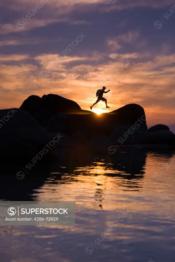  A man jumping on a rock on the shore of Lake Tahoe in California at sunset with reflecting water