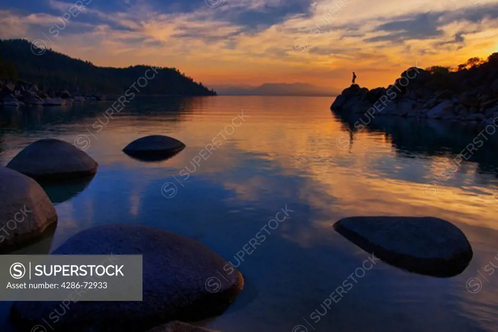 A man standing on a rock on the shore of Lake Tahoe in California at sunset with reflecting water