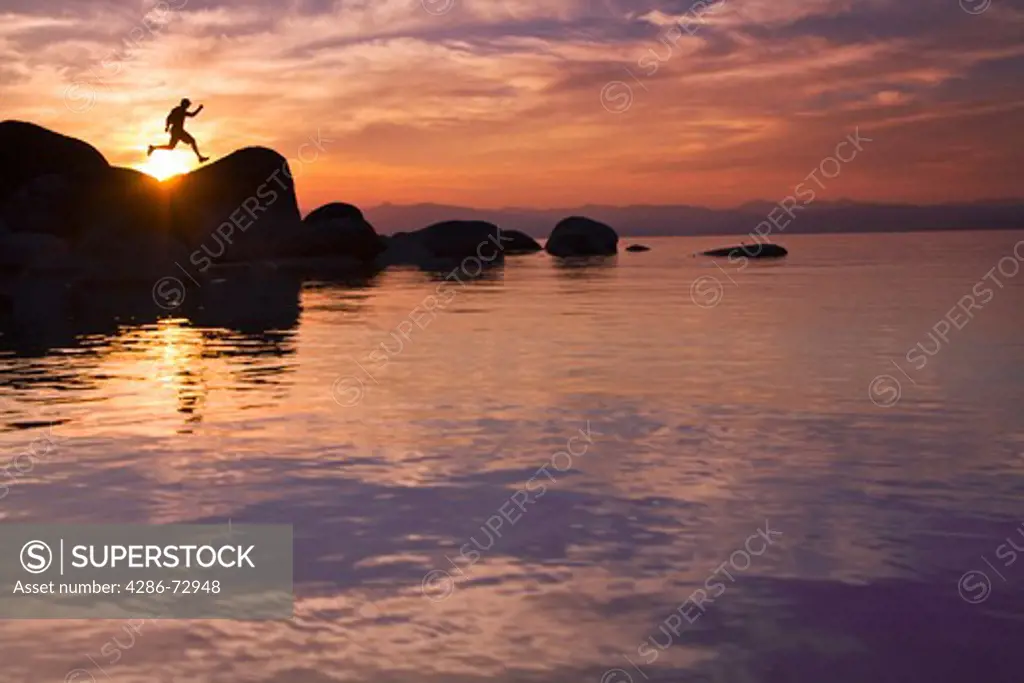  A man jumping on a rock on the shore of Lake Tahoe in California at sunset with reflecting water