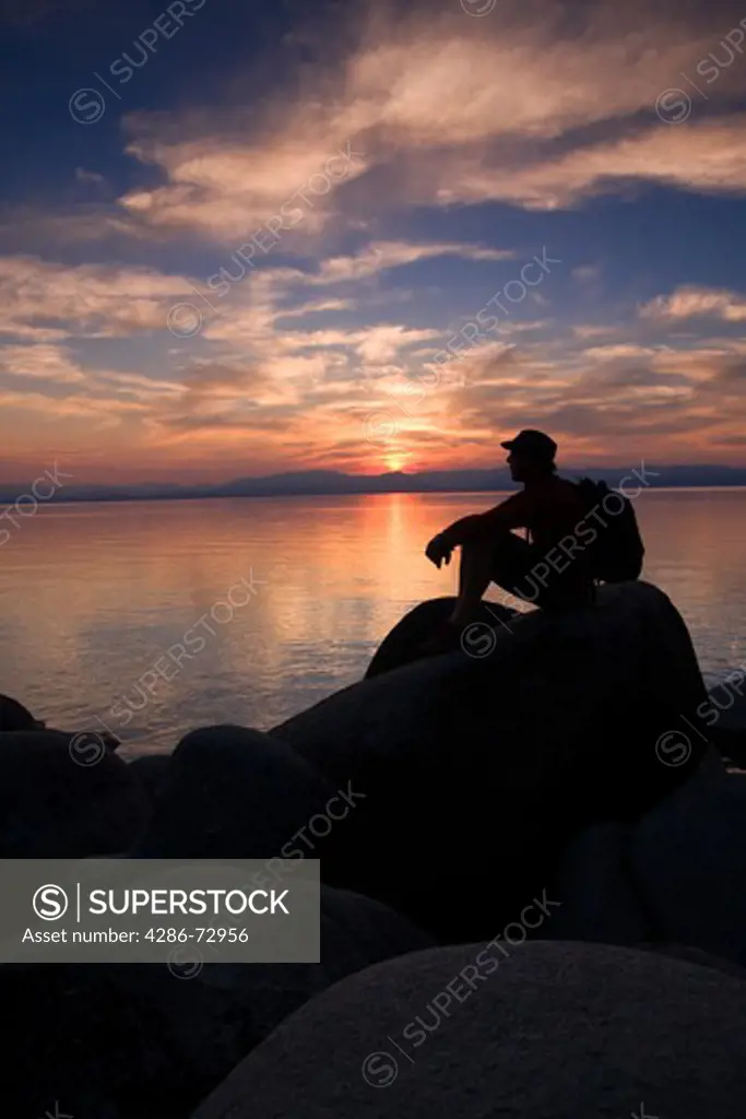  A man sitting on a rock on the shore of Lake Tahoe in California at sunset with reflecting water