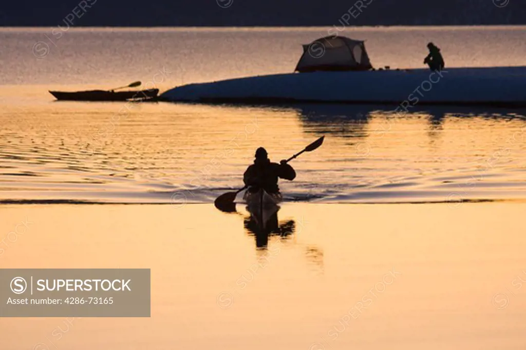 A man and his reflection kayaking past a woman and a tent at sunrise in winter on Lake Tahoe California