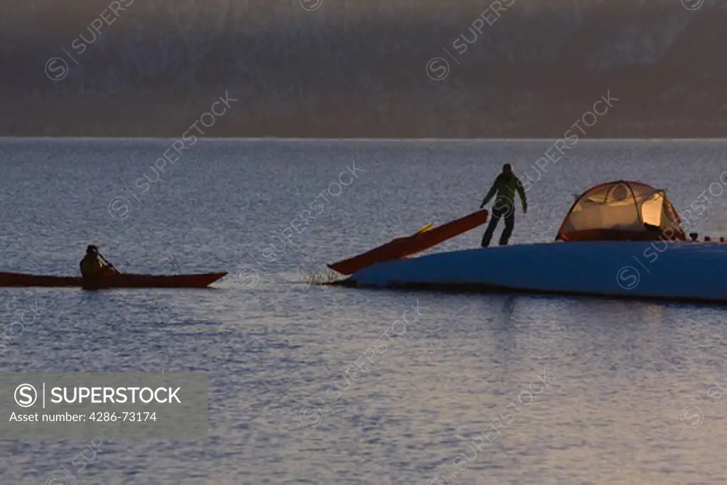 A man sea kayaking past a woman camping on the snowy shore of Lake Tahoe in California in winter.