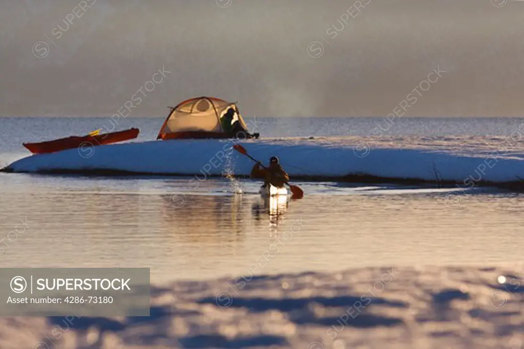 A man sea kayaking past a woman camping on the snowy shore of Lake Tahoe in California in winter.