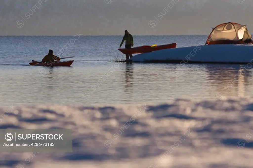 A man in a sea kayak paddling along the shore near a woman and a tent on Lake Tahoe in California on a snowy day at sunrise