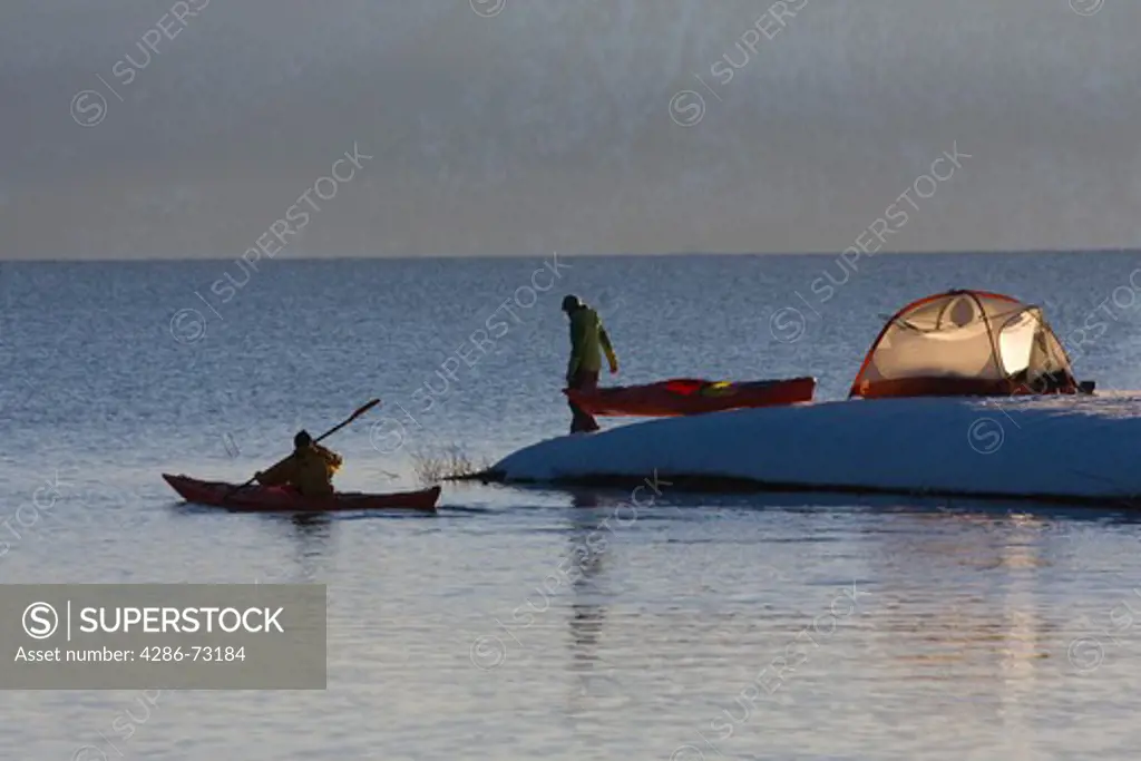 A man in a sea kayak paddling along the shore near a woman and a tent on Lake Tahoe in California on a snowy day at sunrise