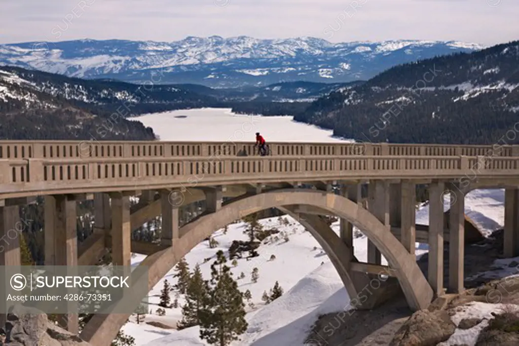  A man climbing uphill on a bicyle over a bridge in winter on Donner Summit in California