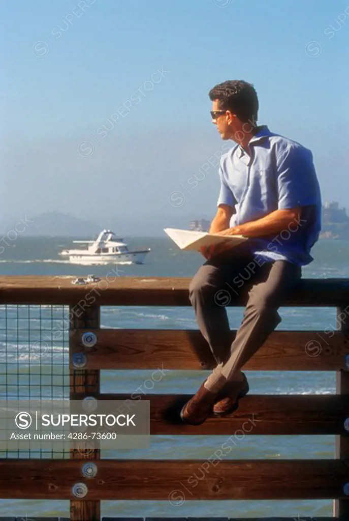 A man sitting on a fence overlooking San Francisco Bay reading a book in California