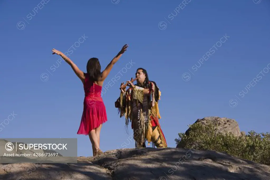 A Lakota Native American Indian warrior in traditional dress playing a love song on a flute to a dancing young maiden on Donner Summit near Truckee in California