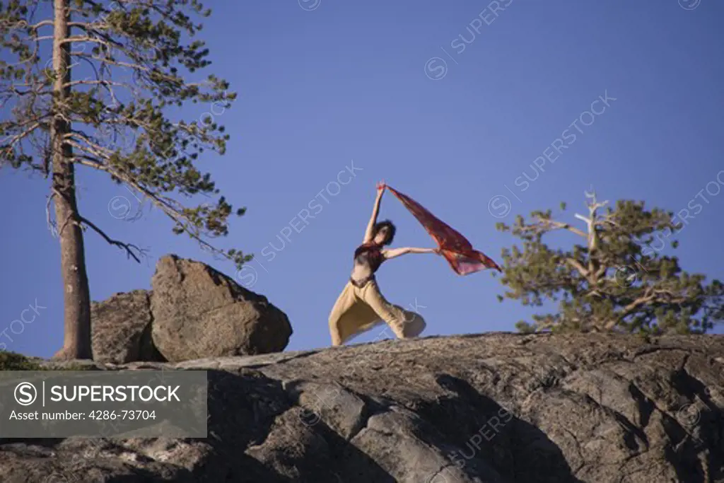 A dancer with a blowing scarf on top of a rock next to boulders and pine trees in the Sierra mountains near Truckee California